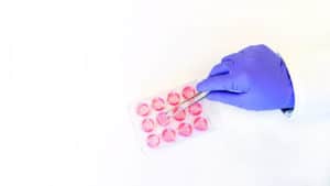 A lab worker's hand in a purple glove holding metal forceps to pick up a tissue insert from a 24-well plate.